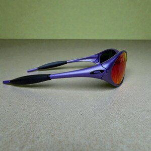 May include: Purple sunglasses with a black logo on the frame and black earpieces. The lenses are a gradient of red and orange. The sunglasses are angled, showing the shape of the frame and lenses.