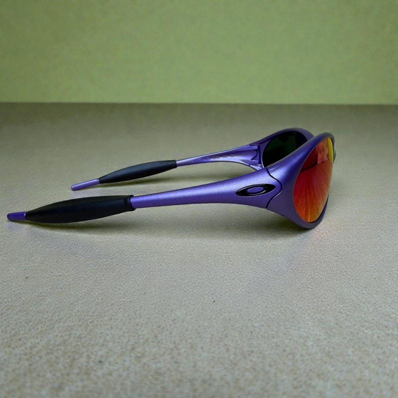 May include: Purple sunglasses with a black logo on the frame and black earpieces. The lenses are a gradient of red and orange. The sunglasses are angled, showing the shape of the frame and lenses.