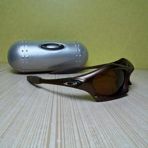 May include: Brown sunglasses with a matching case. The sunglasses have a curved frame and tinted lenses. The case is silver with a riveted design. The sunglasses are resting on a light-colored wooden surface.