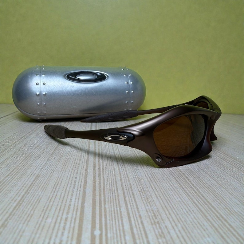 May include: Brown sunglasses with a matching case. The sunglasses have a curved frame and tinted lenses. The case is silver with a riveted design. The sunglasses are resting on a light-colored wooden surface.