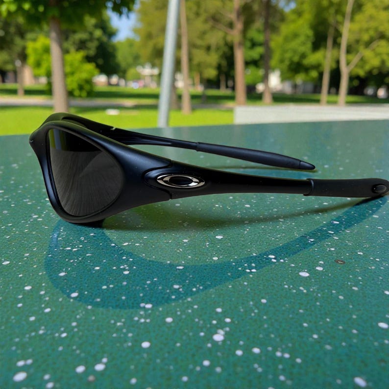 K&ouml;nnte beinhalten: Schwarze Sonnenbrille mit dunklen Gl&auml;sern. Die Sonnenbrille hat ein schlankes, geschwungenes Design mit matter Oberfl&auml;che. Die B&uuml;gel haben ein silbernes Logo. Die Sonnenbrille liegt auf einer gr&uuml;n gesprenkelten Oberfl&auml;che.