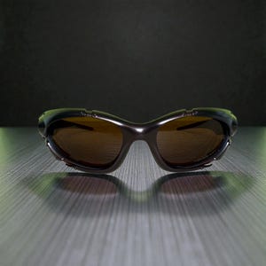 May include: Dark brown sunglasses with a curved frame and brown tinted lenses. The sunglasses have a metallic finish and small rivets along the top of the frame. The lenses are oval-shaped and the bridge is a smooth curve.