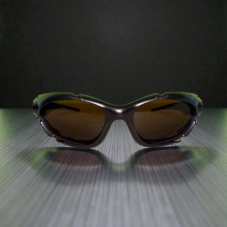 May include: Dark brown sunglasses with a curved frame and brown tinted lenses. The sunglasses have a metallic finish and small rivets along the top of the frame. The lenses are oval-shaped and the bridge is a smooth curve.