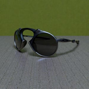 May include: Oakley sunglasses with a silver frame and dark lenses. The sunglasses have a distinctive design with a curved frame and a small, round lens on the side. The lenses are dark, and the frame is a metallic silver colour.