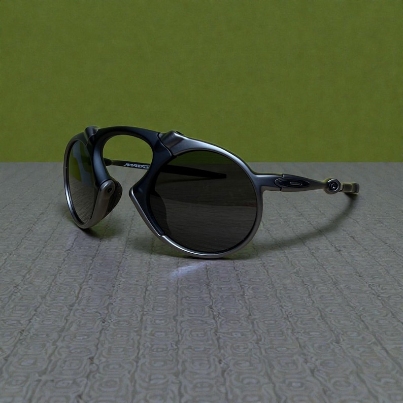 May include: Oakley sunglasses with a silver frame and dark lenses. The sunglasses have a distinctive design with a curved frame and a small, round lens on the side. The lenses are dark, and the frame is a metallic silver colour.