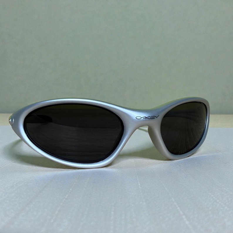 K&ouml;nnte beinhalten: Silberfarbene Sonnenbrille mit dunklen Gl&auml;sern. Die Sonnenbrille hat ein geschwungenes Design und der Markenname "OAKLEY" ist auf dem rechten B&uuml;gel aufgedruckt. Die Sonnenbrille liegt auf einer hellen Oberfl&auml;che.