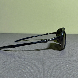 May include: Black sunglasses with a sleek, modern design. The frames are dark, with a matte finish, and the lenses are tinted. The arms are thin and extend back, with a subtle curve. The sunglasses are resting on a grey surface.