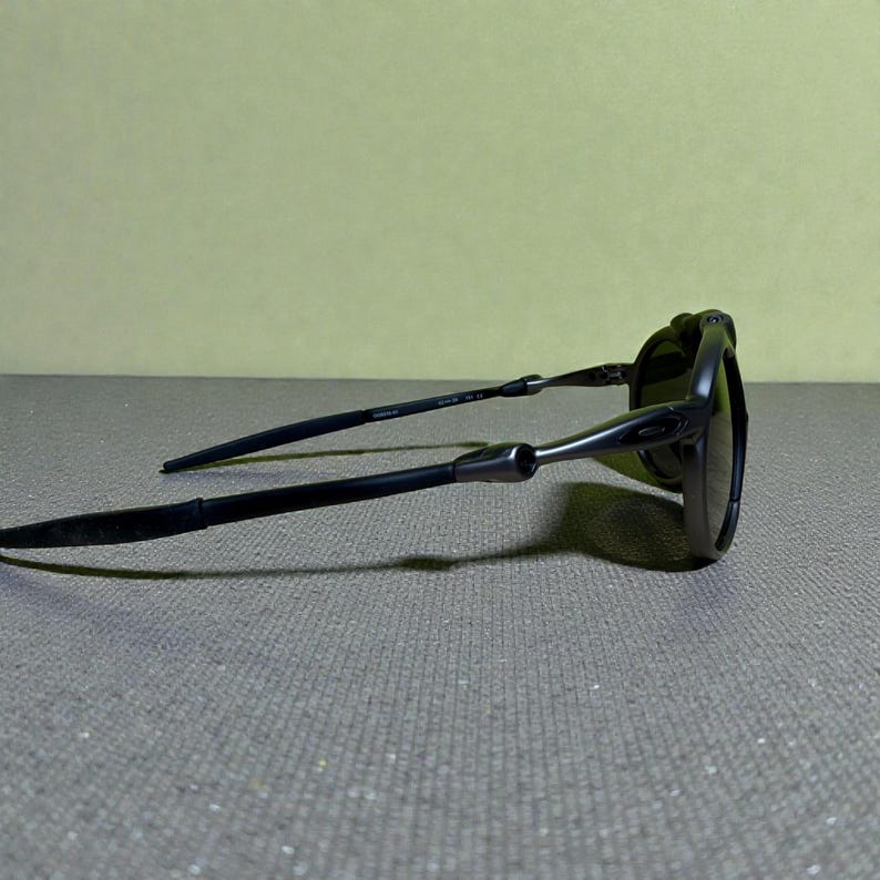 May include: Black sunglasses with a sleek, modern design. The frames are dark, with a matte finish, and the lenses are tinted. The arms are thin and extend back, with a subtle curve. The sunglasses are resting on a grey surface.