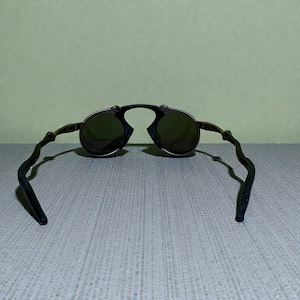 May include: Black round sunglasses with dark lenses. The sunglasses have a unique frame design with curved temples. The lenses are a dark green colour, and the frame appears to be made of a dark metal.