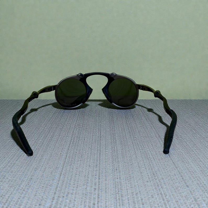 May include: Black round sunglasses with dark lenses. The sunglasses have a unique frame design with curved temples. The lenses are a dark green colour, and the frame appears to be made of a dark metal.