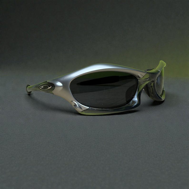 May include: Silver-framed sunglasses with dark lenses. The sunglasses have a sleek, curved design with a prominent brow bar and temples. The Oakley logo is visible on the temple. The overall aesthetic is modern and sporty.