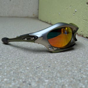 May include: Silver and olive green sports sunglasses with mirrored orange and yellow lenses. The sunglasses have a sleek, aerodynamic design with a curved frame and a small logo on the temple.