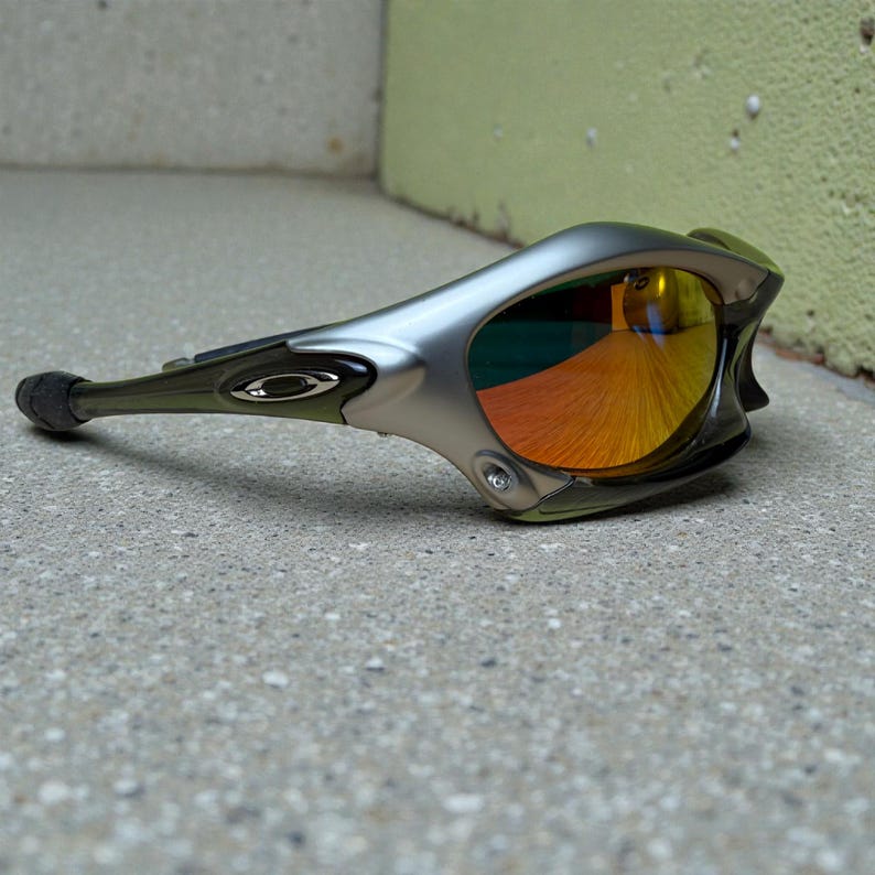 May include: Silver and olive green sports sunglasses with mirrored orange and yellow lenses. The sunglasses have a sleek, aerodynamic design with a curved frame and a small logo on the temple.