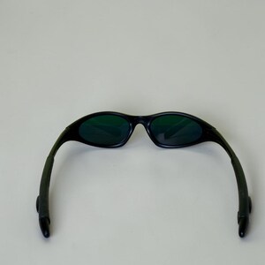 May include: Black sunglasses with green tinted lenses. The sunglasses have a curved frame and arms, and are set against a plain background. The lenses are oval-shaped.