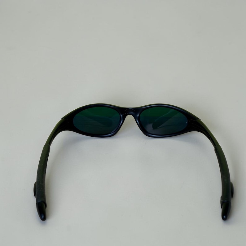 May include: Black sunglasses with green tinted lenses. The sunglasses have a curved frame and arms, and are set against a plain background. The lenses are oval-shaped.