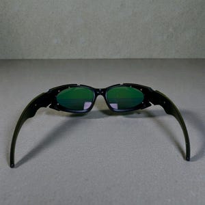 Może przedstawiać: Czarne okulary przeciwsłoneczne z zielonymi soczewkami. Okulary przeciwsłoneczne mają elegancki, nowoczesny design z zakrzywioną oprawką i zausznikami. Soczewki są w żywym zielonym kolorze, a oprawka jest czarna. Okulary przeciwsłoneczne mają chronić oczy przed słońcem.