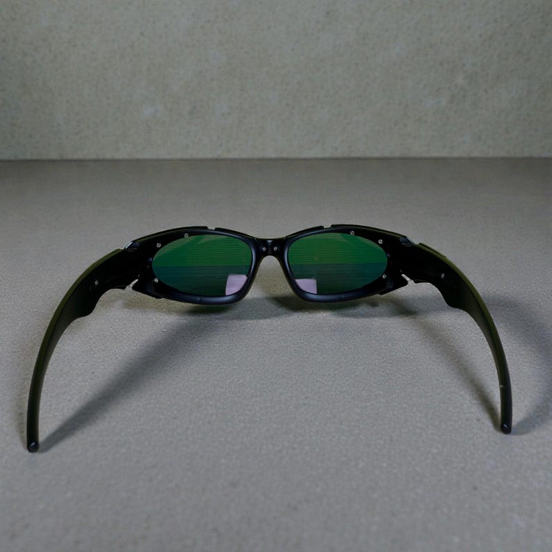 Może przedstawiać: Czarne okulary przeciwsłoneczne z zielonymi soczewkami. Okulary przeciwsłoneczne mają elegancki, nowoczesny design z zakrzywioną oprawką i zausznikami. Soczewki są w żywym zielonym kolorze, a oprawka jest czarna. Okulary przeciwsłoneczne mają chronić oczy przed słońcem.