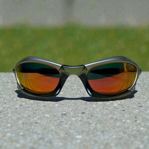 May include: Sport sunglasses with a dark gray frame and mirrored orange lenses. The lenses reflect a vibrant orange hue, contrasting with the darker frame. The sunglasses are designed for sports and outdoor activities.