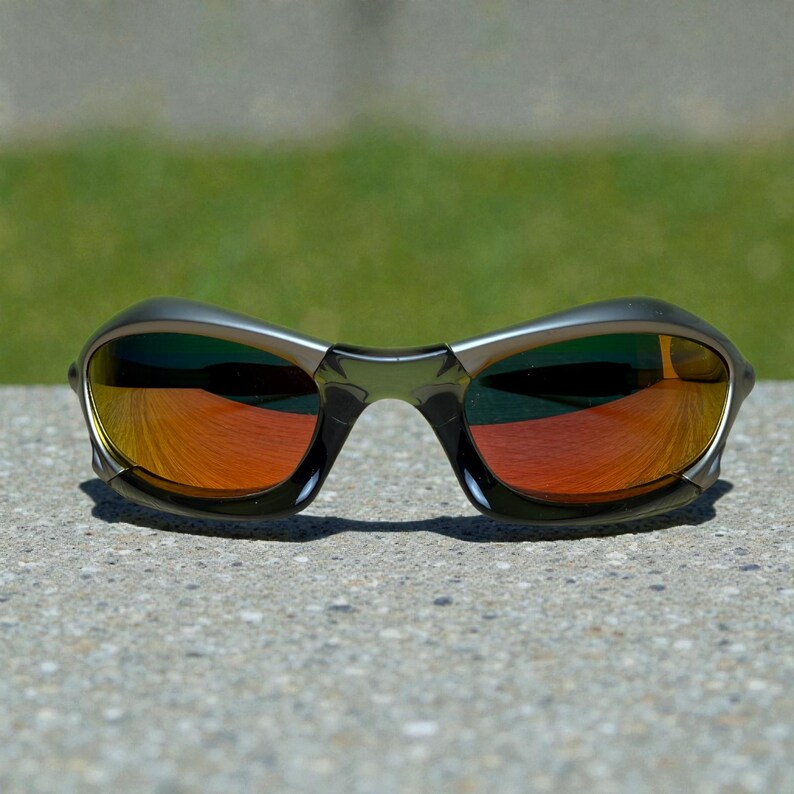 May include: Sport sunglasses with a dark gray frame and mirrored orange lenses. The lenses reflect a vibrant orange hue, contrasting with the darker frame. The sunglasses are designed for sports and outdoor activities.