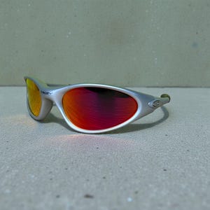 Op de afbeelding: Zilverkleurige zonnebril met rode en gele glazen. De zonnebril heeft een strak, modern design met een gebogen montuur en een klein logo aan de zijkant. De glazen lopen over van rood naar geel.