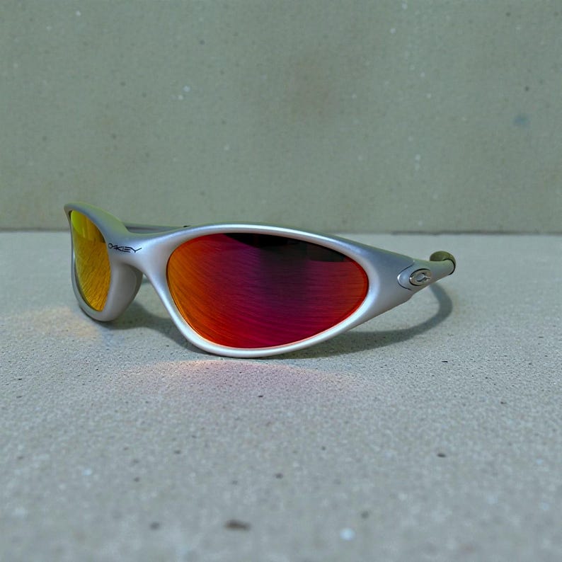 Op de afbeelding: Zilverkleurige zonnebril met rode en gele glazen. De zonnebril heeft een strak, modern design met een gebogen montuur en een klein logo aan de zijkant. De glazen lopen over van rood naar geel.