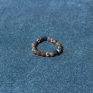 Könnte beinhalten: Ein kreisförmiger Ring aus kleinen, facettierten, mehrfarbigen Edelsteinperlen, die mit winzigen, reflektierenden goldfarbenen Metallkugeln durchsetzt sind. Die Perlen zeigen eine Vielzahl von erdigen und juwelenartigen Tönen in Rot, Blau und Braun.
