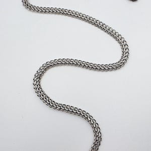 May include: A silver-colored necklace with a woven chain design. The necklace is displayed on a white background. The chain is made of small, interconnected links, creating a textured appearance. The necklace has a clasp closure.