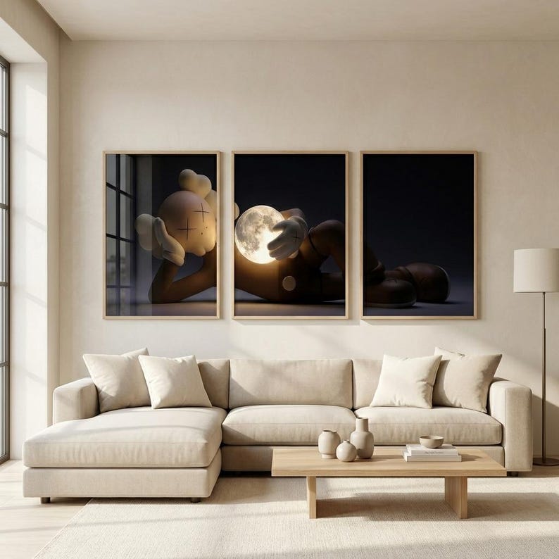 May include: A triptych of framed wall art prints featuring a brown cartoon character with X-shaped eyes lying down and holding a glowing, luminous full moon. The artwork is displayed in three separate light-colored wooden frames above a beige sofa.