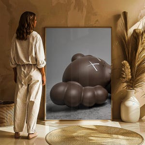 May include: A framed artwork featuring a large, brown, cartoon-like bear figure with white 'X' marks for eyes. The bear is lying down. The artwork is in a room with a woman standing nearby.