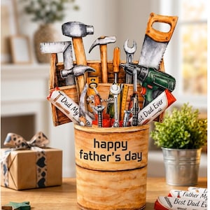 Puede incluir: Un recipiente de madera con el texto "happy father's day" y una colección de herramientas, incluyendo martillos, una sierra y destornilladores. El recipiente está sobre una mesa de madera, con una caja de regalo envuelta y una pequeña planta en el fondo.