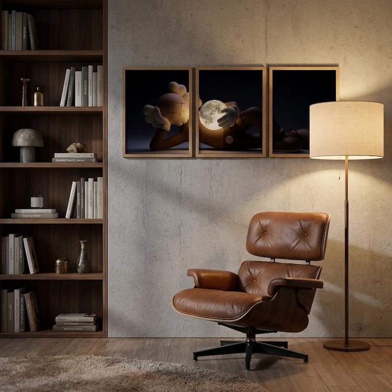 May include: A brown leather lounge chair with a tufted back and seat, positioned next to a tall floor lamp with a beige drum shade. Three framed prints depicting a cartoon character holding a glowing moon are mounted on the wall above the chair.
