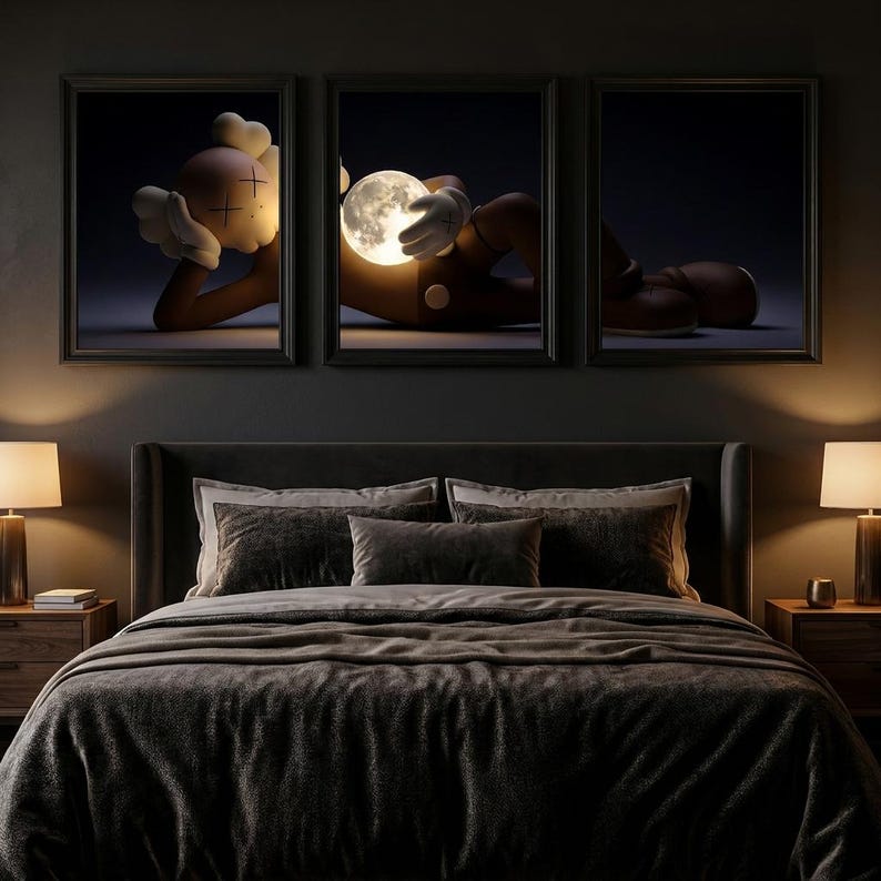 May include: A triptych of framed wall art depicting a brown cartoon character with X-shaped eyes resting and holding a glowing, luminous moon. The artwork is displayed above a dark-colored bed with matching bedside lamps on either side.