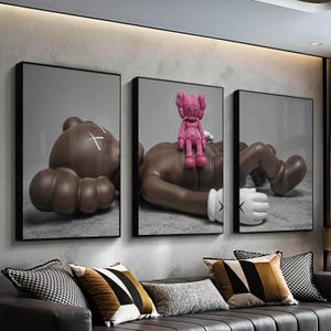 May include: Three framed art prints featuring a brown figure with a white cross, a brown figure with a pink figure on top, and a brown figure with a white glove. The art is displayed above a sofa with decorative pillows.