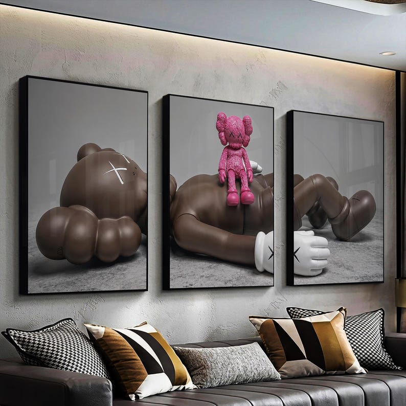 May include: Three framed art prints featuring a brown figure with a white cross, a brown figure with a pink figure on top, and a brown figure with a white glove. The art is displayed above a sofa with decorative pillows.