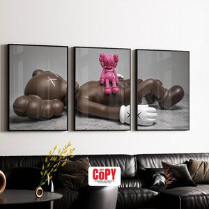 May include: Triptych wall art featuring three framed prints. Each print depicts a stylized brown figure in a relaxed pose. A pink figure is perched on one of the brown figures. The prints are displayed above a black leather sofa.