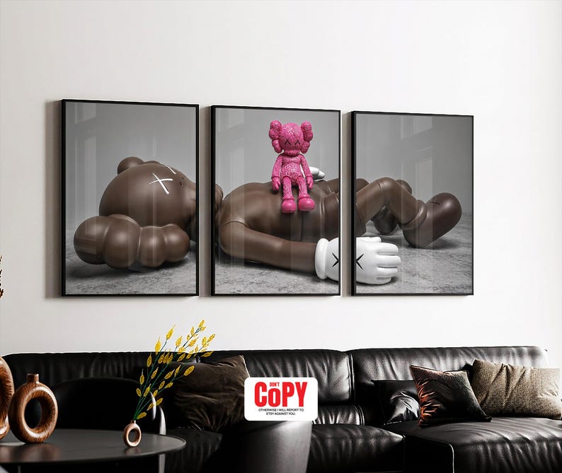 May include: Triptych wall art featuring three framed prints. Each print depicts a stylized brown figure in a relaxed pose. A pink figure is perched on one of the brown figures. The prints are displayed above a black leather sofa.