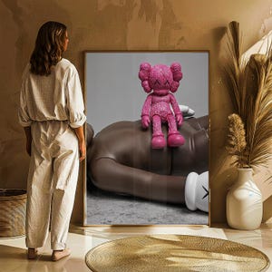 May include: A framed artwork featuring a pink KAWS Companion figure atop a large, brown figure. The artwork is displayed in a room with a woman standing nearby. The room has a neutral color palette with a gold-toned rug and a white vase.