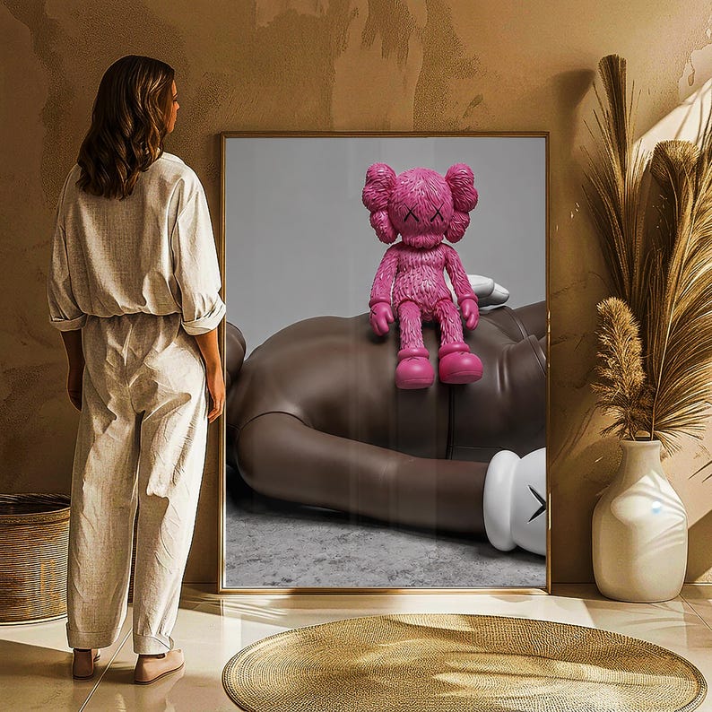 May include: A framed artwork featuring a pink KAWS Companion figure atop a large, brown figure. The artwork is displayed in a room with a woman standing nearby. The room has a neutral color palette with a gold-toned rug and a white vase.