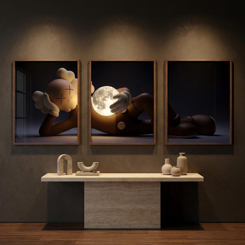 May include: A triptych of framed wall art featuring a brown cartoon character with X-shaped eyes lying down and holding a glowing, cratered moon. The artwork is displayed above a light beige stone console table with small decorative ceramic vases.