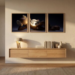 May include: A triptych of three framed prints featuring a brown cartoon character with X-shaped eyes lying down and holding a glowing, detailed moon. The artwork is displayed above a light oak floating console table with books and a lamp.