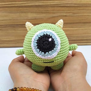 May include: A handmade, green crocheted monster with one large eye, small horns, and tiny arms and legs. The eye is black with a white highlight, surrounded by a light blue and white ring. The monster is held in a person's hands.