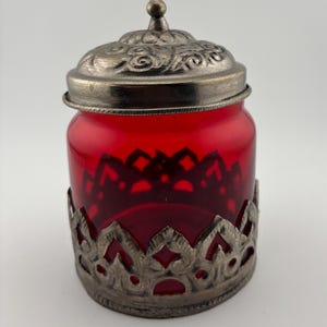 May include: A small, cylindrical red glass jar featuring a removable silver-toned metal lid with an ornate, embossed swirl pattern. The base of the jar is encased in a decorative silver-toned metal sleeve with a repeating cutout arch design.