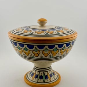 May include: A ceramic lidded bowl with a pedestal base, featuring a cream-coloured background decorated with hand-painted patterns in vibrant yellow, deep blue, and soft green. The lid has a small knob handle and matching floral motifs.
