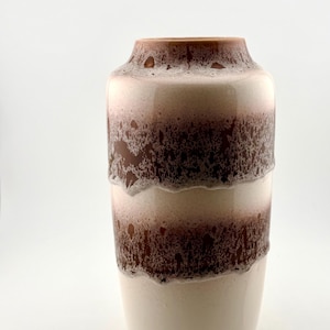 May include: A cylindrical ceramic vase featuring a smooth cream-coloured base and two distinct horizontal bands of textured, dark brown glaze. The glaze has a mottled, drip-like appearance that contrasts with the solid, light-coloured sections.