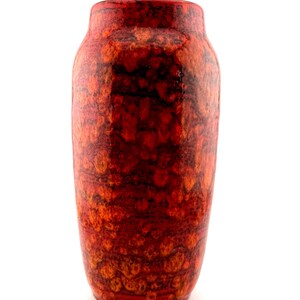 May include: A cylindrical ceramic vase featuring a vibrant red and orange mottled glaze with dark brown accents. The surface has a textured, volcanic appearance with a glossy finish, a narrow neck, and a rounded base, standing upright.