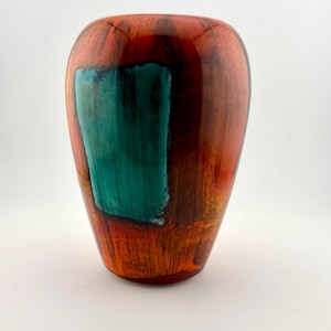 May include: A smooth, rounded wooden vase with a polished finish. The exterior features a rich, warm reddish-brown wood grain pattern contrasted by a prominent, rectangular patch of vibrant teal colour applied to the side of the vessel.