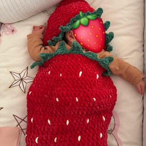 May include: A red crocheted strawberry-shaped sleep sack with a green crocheted leaf collar and a matching hood. The red body features small white yarn accents resembling strawberry seeds. The garment is made of a soft, plush yarn texture.