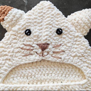 May include: A cream-colored crocheted hood featuring a cat-like face with two pointed ears, one of which has a brown tip. The face includes embroidered black eyes, a brown triangular nose, and brown whiskers stitched onto the textured yarn surface.
