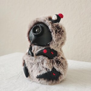 May include: A plush creature covered in brown and white faux fur, featuring a black leather-like mask with a single circular eye lens. It has black leather-like appendages with red stitching and small red floral accents, plus a tiny skull topper.