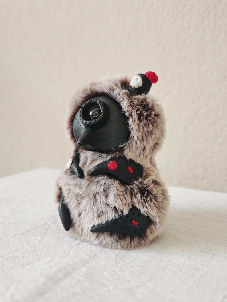 May include: A plush creature covered in brown and white faux fur, featuring a black leather-like mask with a single circular eye lens. It has black leather-like appendages with red stitching and small red floral accents, plus a tiny skull topper.